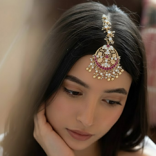 Gold-plated maang tikka with red and blue stones, pearl beads, and intricate floral design for bridal and festive wear.
