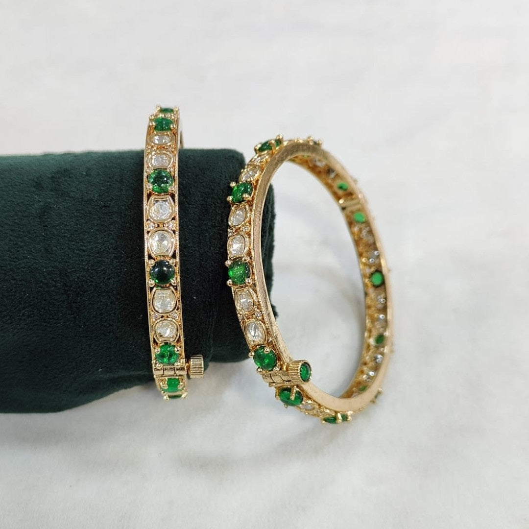 Set of gold-plated bangles adorned with alternating green and white Kundan stones, displayed on a dark green velvet roll against a white background.