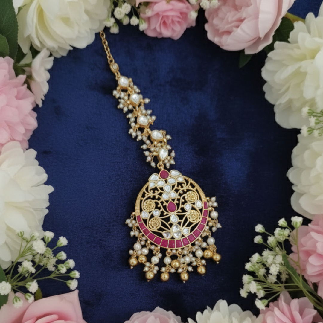 Gold-plated maang tikka with red and blue stones, pearl beads, and intricate floral design for bridal and festive wear.