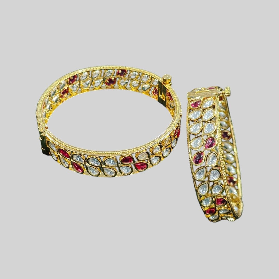 Pair of gold-plated bangles with alternating red and white teardrop-shaped Kundan stones, displayed on dark green velvet cloth with pink roses in the background – traditional bridal kada set.