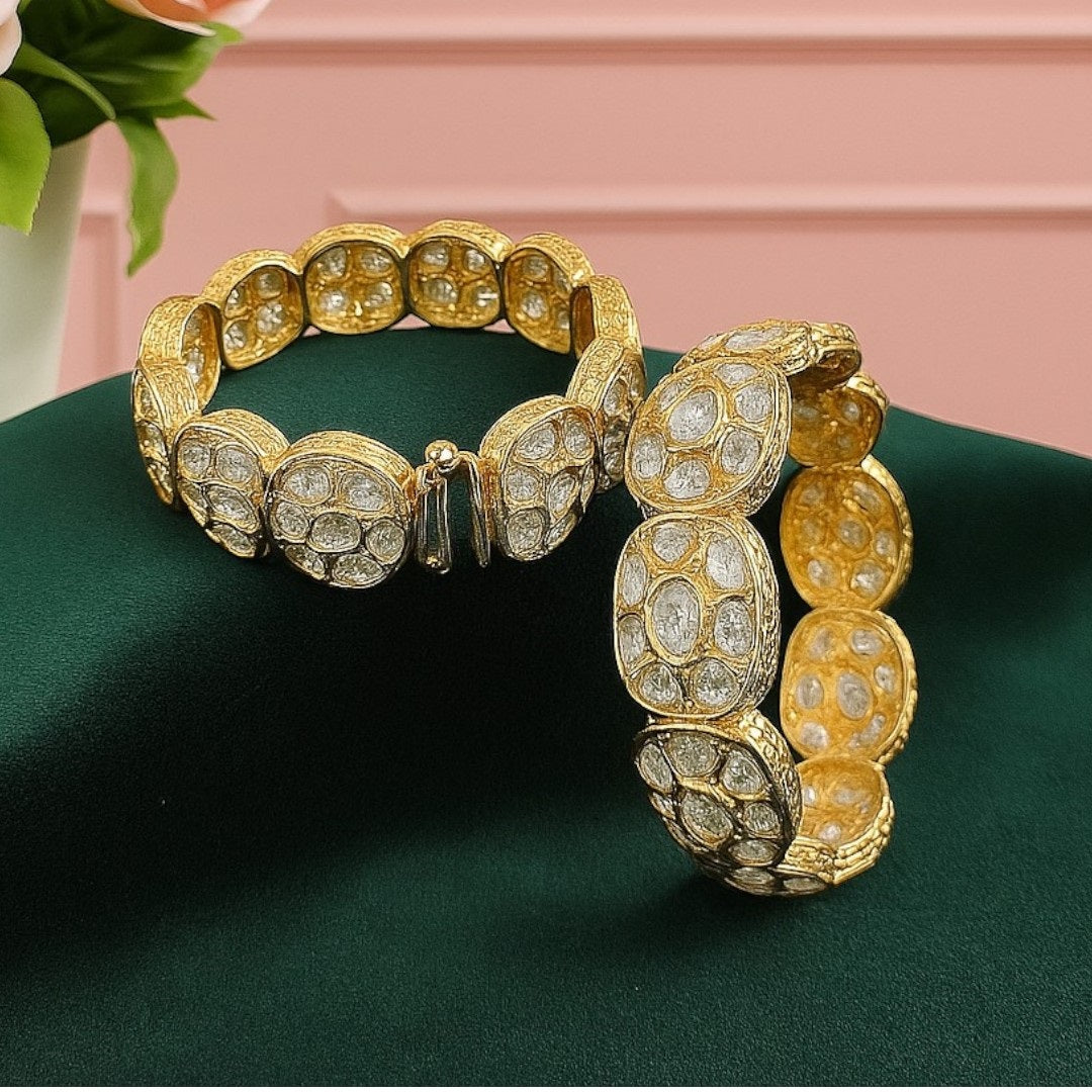 Set of two broad gold-plated bangles featuring oval white Kundan-style stones, displayed on a green velvet cloth with a pink floral background – ethnic bridal kada jewelry.