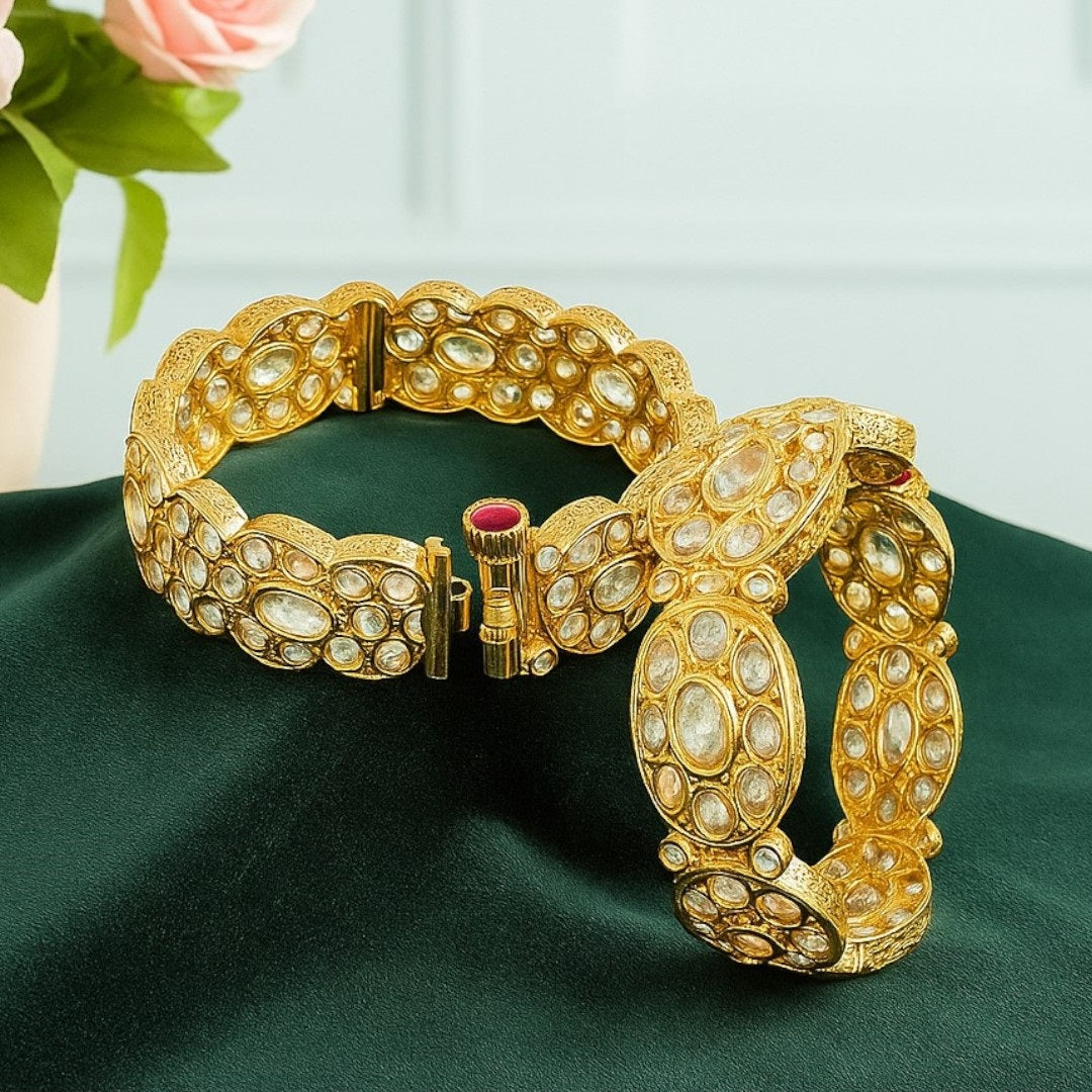 Pair of gold-plated oval Kundan stone bangles with red screw clasp detail, displayed on a green velvet cloth with pink floral background – bridal kada set for women.