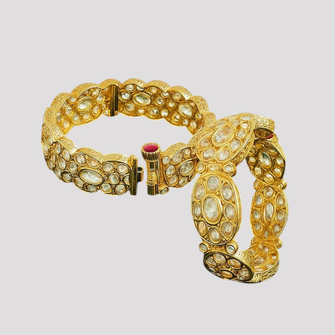 Pair of gold-plated oval Kundan stone bangles with red screw clasp detail, displayed on a green velvet cloth with pink floral background – bridal kada set for women.