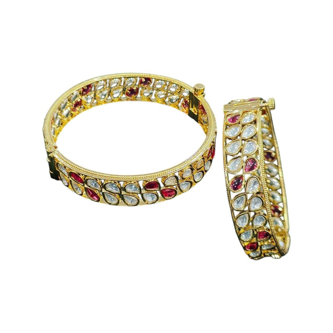 Pair of gold-plated bangles with alternating red and white teardrop-shaped Kundan stones, displayed on dark green velvet cloth with pink roses in the background – traditional bridal kada set.