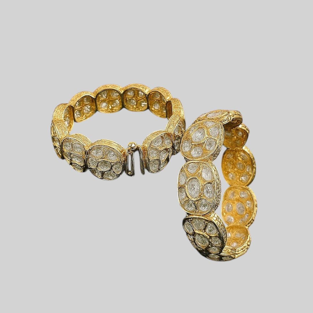 Set of two broad gold-plated bangles featuring oval white Kundan-style stones, displayed on a green velvet cloth with a pink floral background – ethnic bridal kada jewelry.