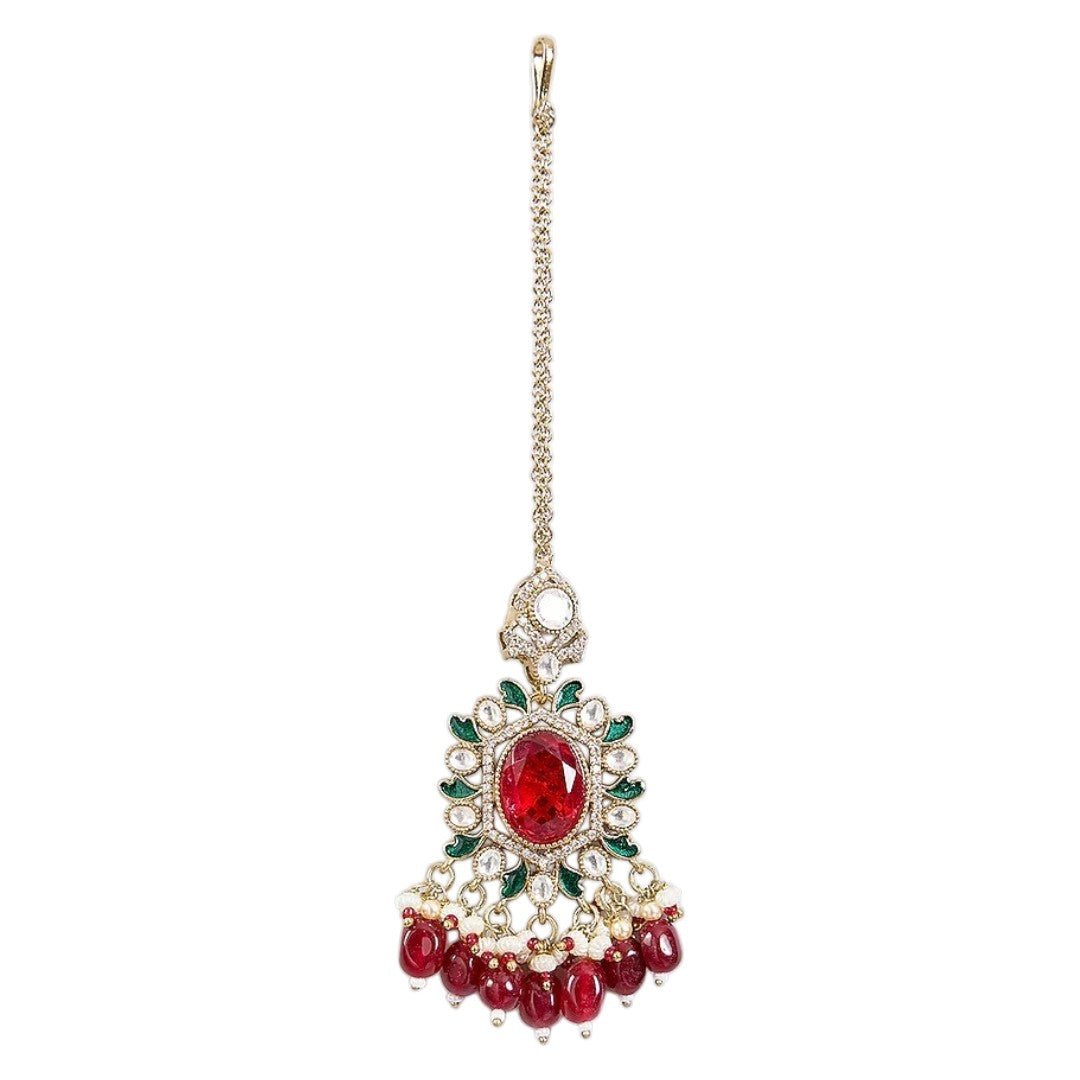 Gold-toned Kundan maang tikka with ruby red oval stone, green enamel detailing, sparkling Kundan stones, and red bead drops for bridal and festive wear.