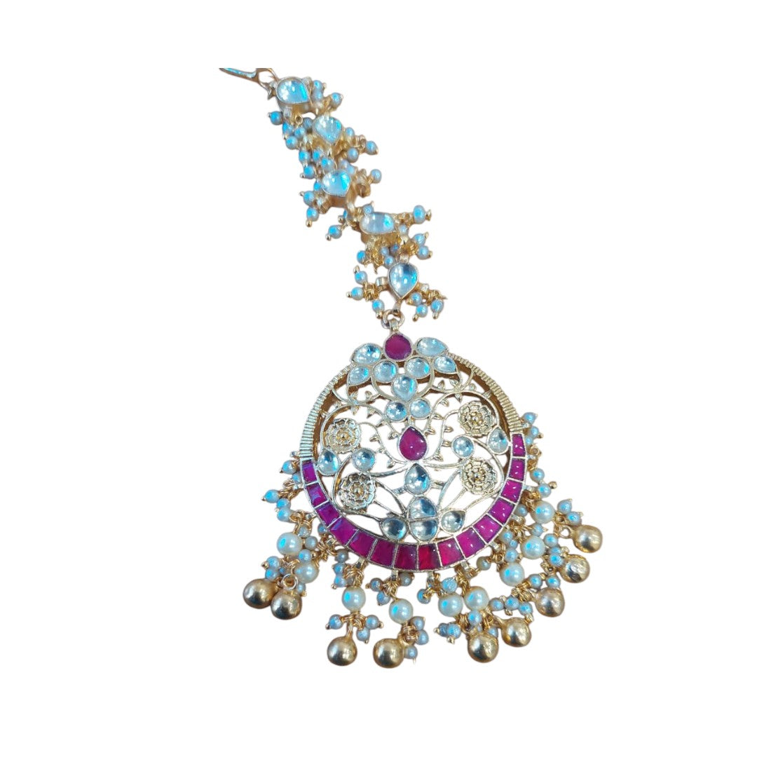Gold-plated maang tikka with red and blue stones, pearl beads, and intricate floral design for bridal and festive wear.