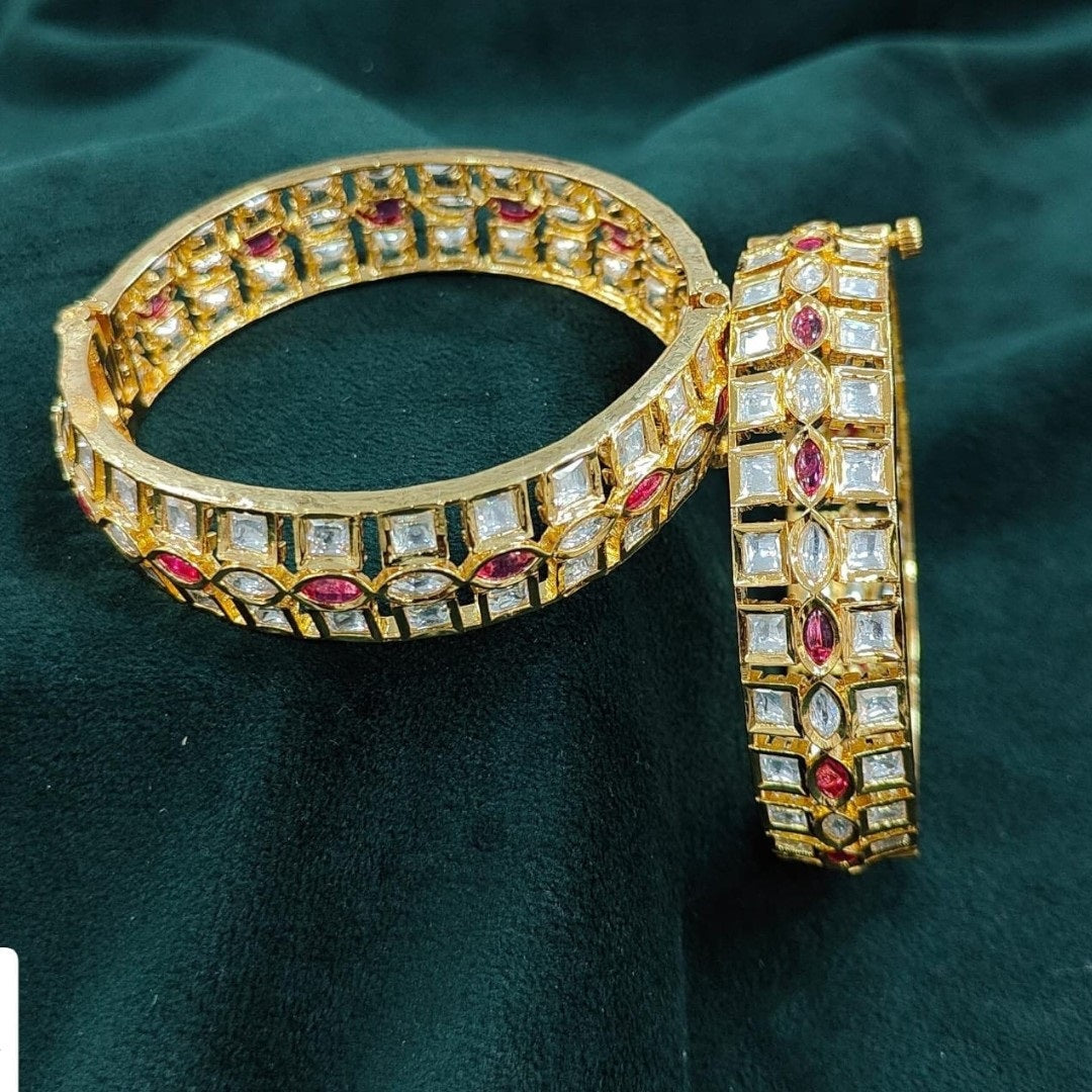 Elegant gold-plated bangles with square white stones and marquise ruby stones, placed on a marble background with yellow daisies – perfect for bridal and festive wear.