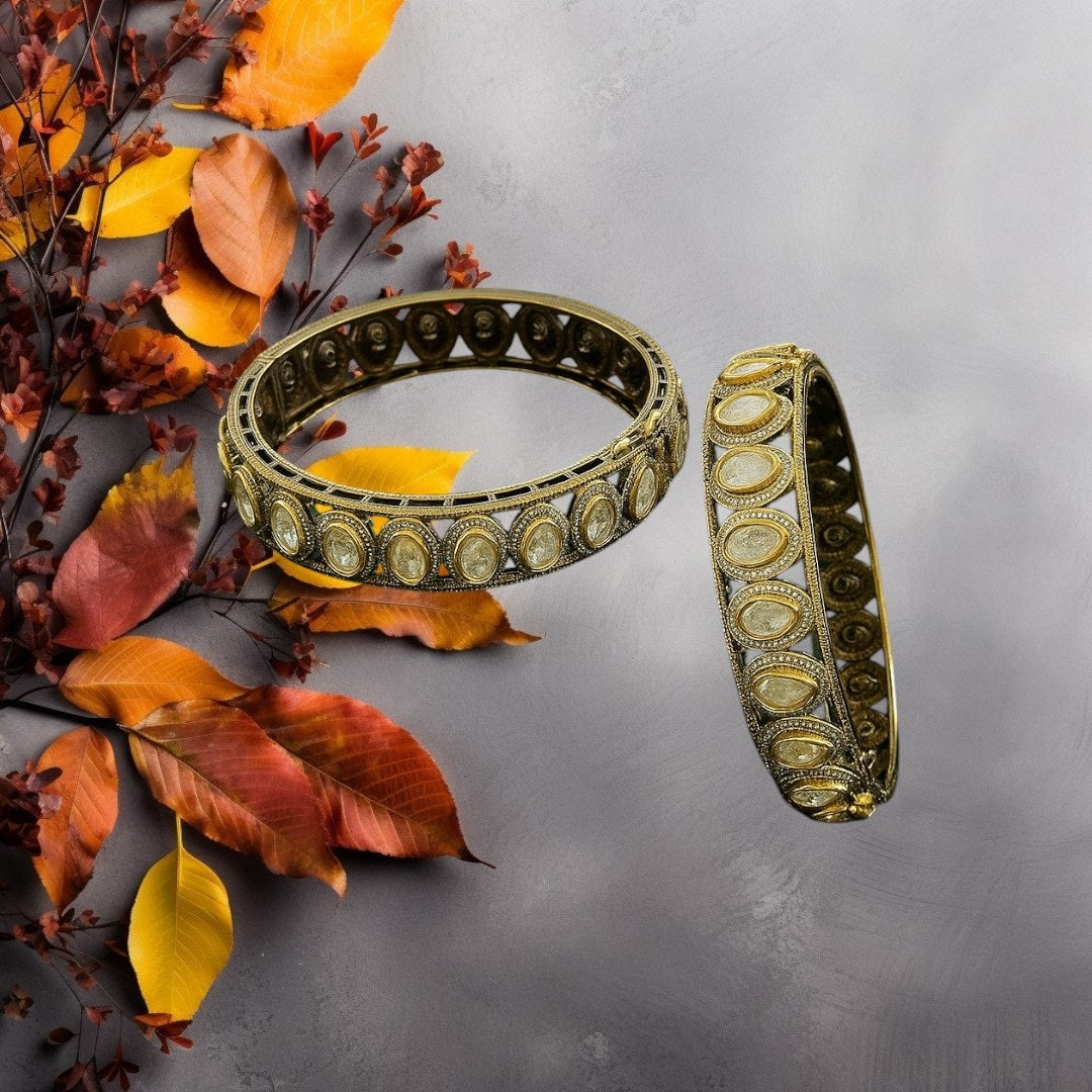"Antique gold-plated bangles with oval polki stones in traditional Kundan style, placed on a green velvet cloth with floral background"