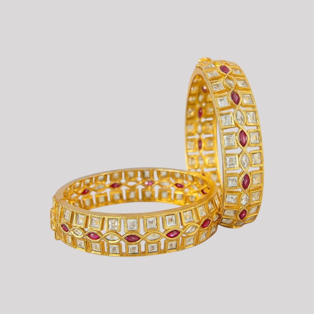 Elegant gold-plated bangles with square white stones and marquise ruby stones, placed on a marble background with yellow daisies – perfect for bridal and festive wear.