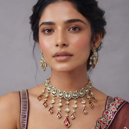 Gold-tone kundan necklace set with red kite-shaped drops, green accent stones and matching chandelier earrings on a grey display bust