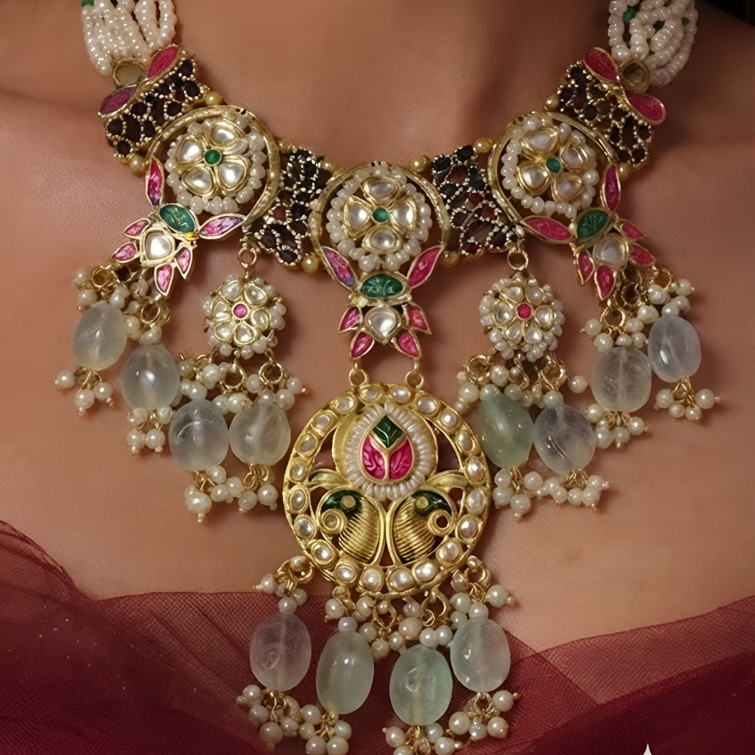 "Antique gold Kundan necklace and earring set with multi-strand pearl beading, ruby-green stone work, peacock detail and green bead drops, traditional jewellery on maroon backdrop"