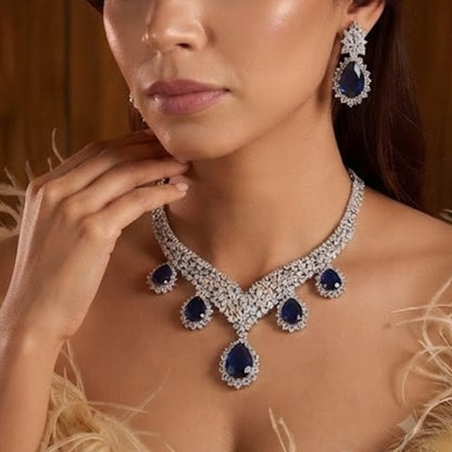"Silver rhodium-plated V-shaped necklace set with white American diamonds and blue sapphire pear-drop stones, paired with matching teardrop earrings"