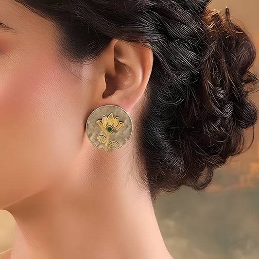 Gold lotus flower stud earrings with green emerald center on hammered oxidized silver disc, placed on a beige fabric background