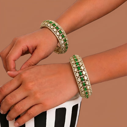 Pair of gold-finish wide kada bangles with emerald-green oval stones, polki-style clear stones, and American diamond edging