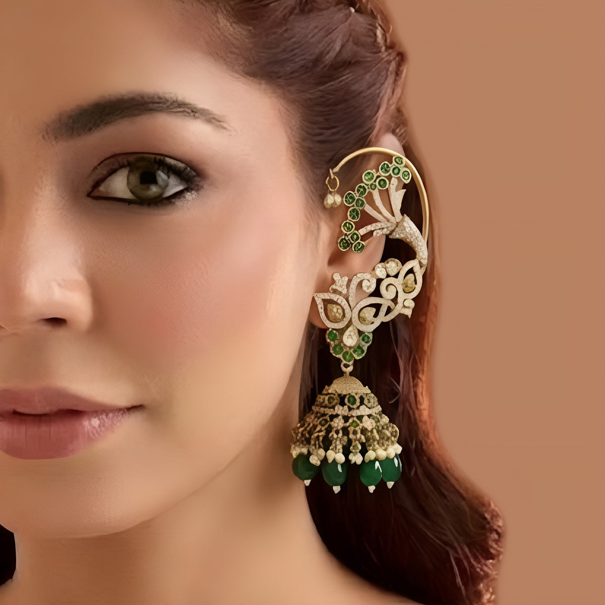 Gold plated emerald green ear cuff jhumka earrings with American diamond stones, green beads and pearl hangings on a white background