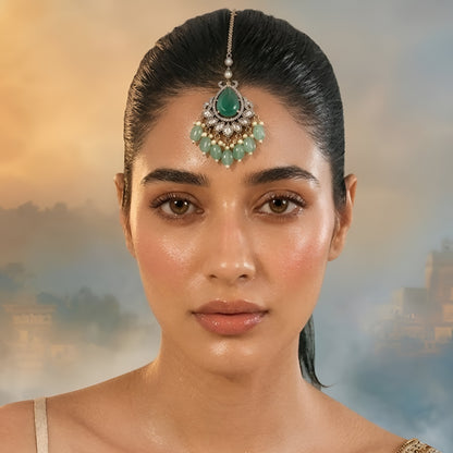 Gold-tone bridal maang tikka featuring a pear-shaped green stone, crystal floral detailing, pearl accents, and green bead drops for traditional Indian wedding jewelry.