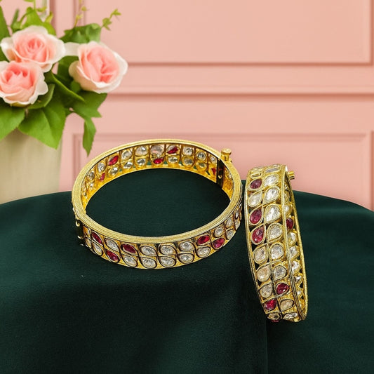 Pair of gold-plated bangles with alternating red and white teardrop-shaped Kundan stones, displayed on dark green velvet cloth with pink roses in the background – traditional bridal kada set.