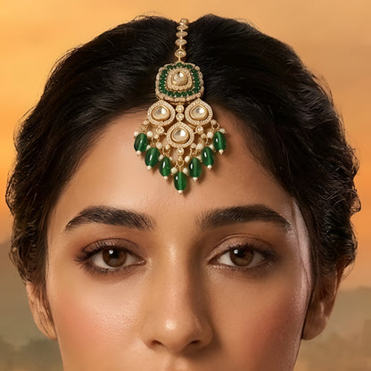 Gold-toned Kundan maang tikka with green stones, teardrop crystals, pearl accents, and dangling green beads for bridal and festive wear.