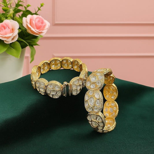 Set of two broad gold-plated bangles featuring oval white Kundan-style stones, displayed on a green velvet cloth with a pink floral background – ethnic bridal kada jewelry.