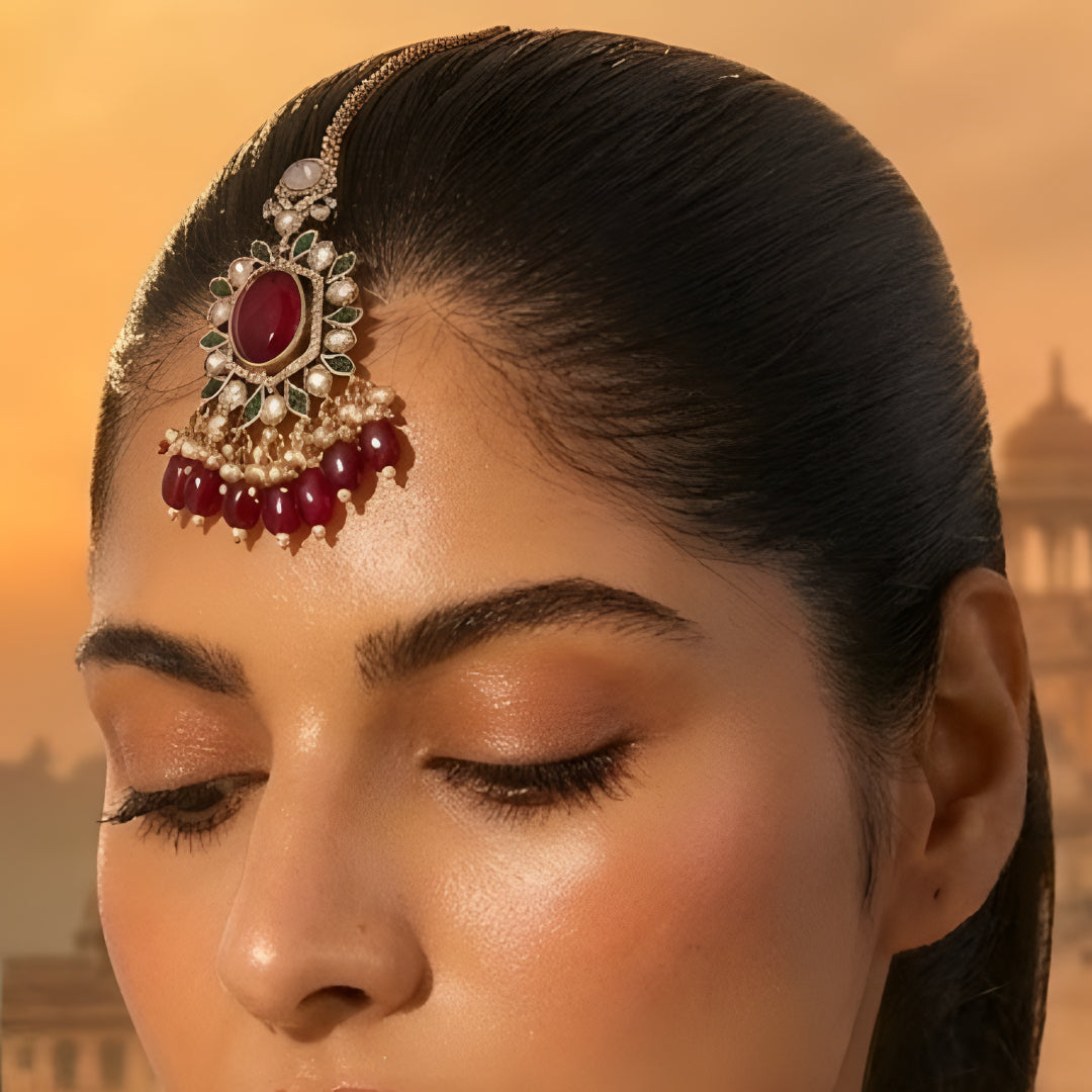 Gold-toned Kundan maang tikka with ruby red oval stone, green enamel detailing, sparkling Kundan stones, and red bead drops for bridal and festive wear.