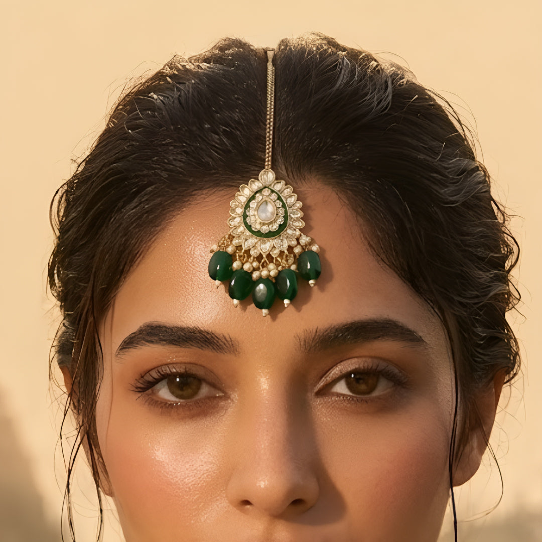 Gold-toned Kundan maang tikka with green enamel detailing, teardrop stones, pearl accents, and emerald-green bead drops for bridal and festive wear.