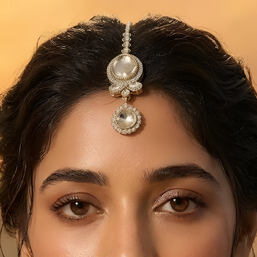 Gold-toned American Diamond oval stone maang tikka with floral crystal design and dangling charm for bridal and festive wear.