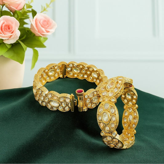 Pair of gold-plated oval Kundan stone bangles with red screw clasp detail, displayed on a green velvet cloth with pink floral background – bridal kada set for women.