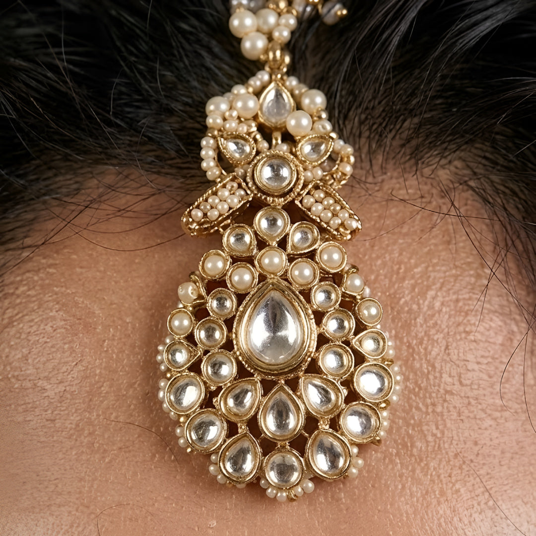 Gold-plated Kundan maang tikka with sparkling clear stones, pearl bead drops, and an elegant bridal design for weddings and festive wear.