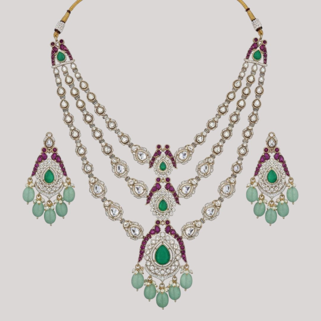 "Triple-layer gold-finish necklace set with green stones, purple detailing, American Diamond accents, pastel green bead drops, and matching earrings displayed on a light background."