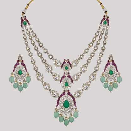 "Triple-layer gold-finish necklace set with green stones, purple detailing, American Diamond accents, pastel green bead drops, and matching earrings displayed on a light background."