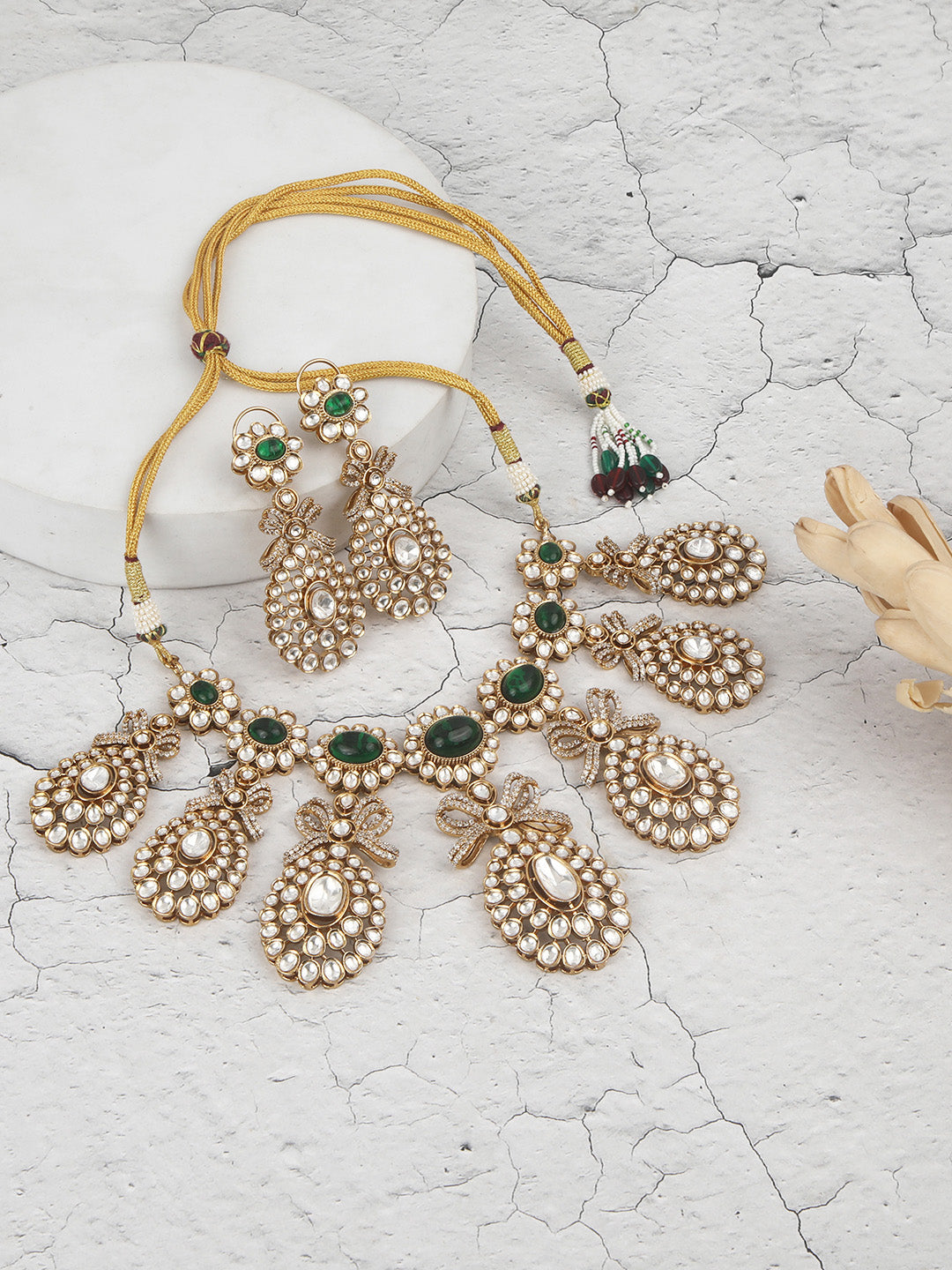 Gold-toned necklace with a braided cord and adjustable tassel tie, featuring intricate white stone detailing with oval green gemstones and multiple dangling teardrop motifs, paired with matching drop earrings, displayed on a plain light background.
