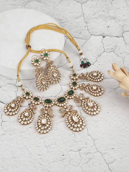 Gold-toned necklace with a braided cord and adjustable tassel tie, featuring intricate white stone detailing with oval green gemstones and multiple dangling teardrop motifs, paired with matching drop earrings, displayed on a plain light background.