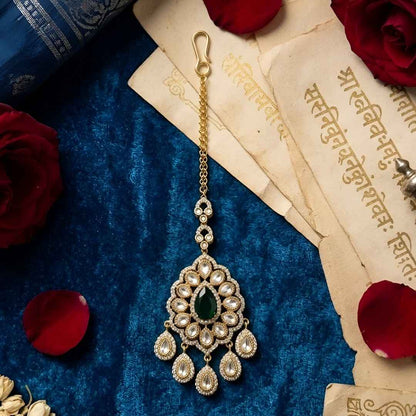 Gold-plated Kundan Maang Tikka with emerald green teardrop stone and white Kundan detailing for bridal and festive wear.