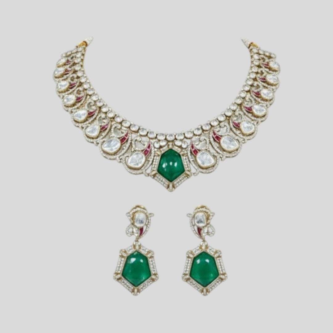 "Gold-finish necklace set with emerald green stones, white American Diamond detailing, red accents, and matching drop earrings displayed on a light background."