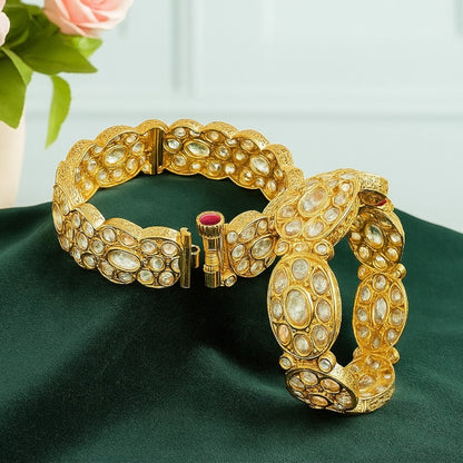 Pair of gold-plated oval Kundan stone bangles with red screw clasp detail, displayed on a green velvet cloth with pink floral background – bridal kada set for women.