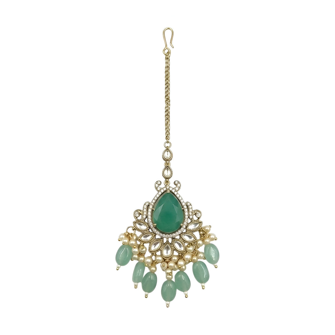 Gold-tone bridal maang tikka featuring a pear-shaped green stone, crystal floral detailing, pearl accents, and green bead drops for traditional Indian wedding jewelry.