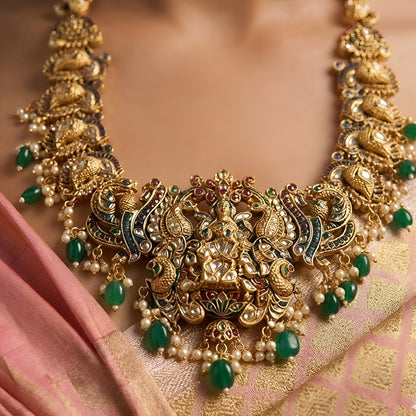 Antique gold Lakshmi temple necklace featuring traditional South Indian craftsmanship, ruby and green stone detailing, intricate deity motifs, and green bead drops for bridal and festive occasions.