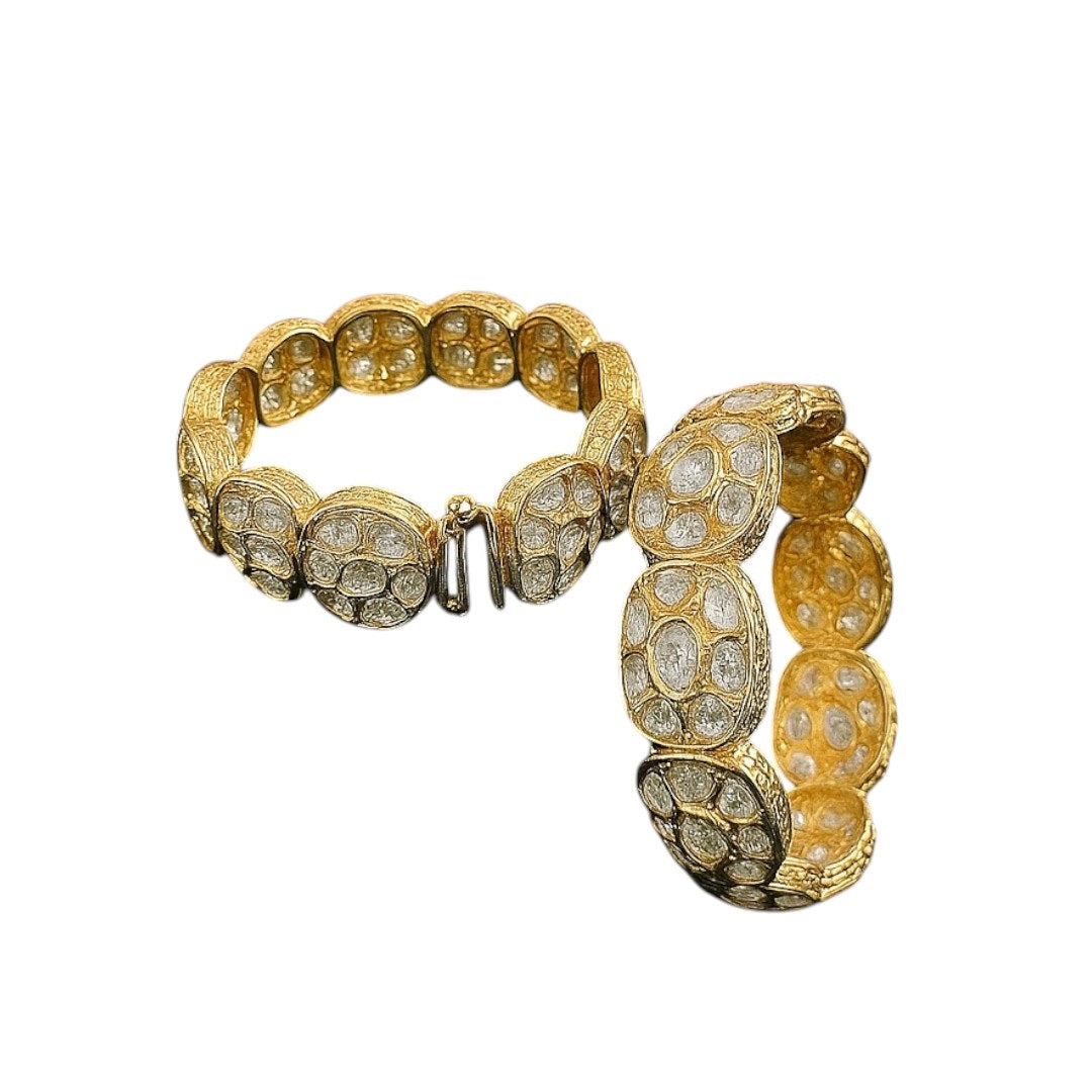 Set of two broad gold-plated bangles featuring oval white Kundan-style stones, displayed on a green velvet cloth with a pink floral background – ethnic bridal kada jewelry.