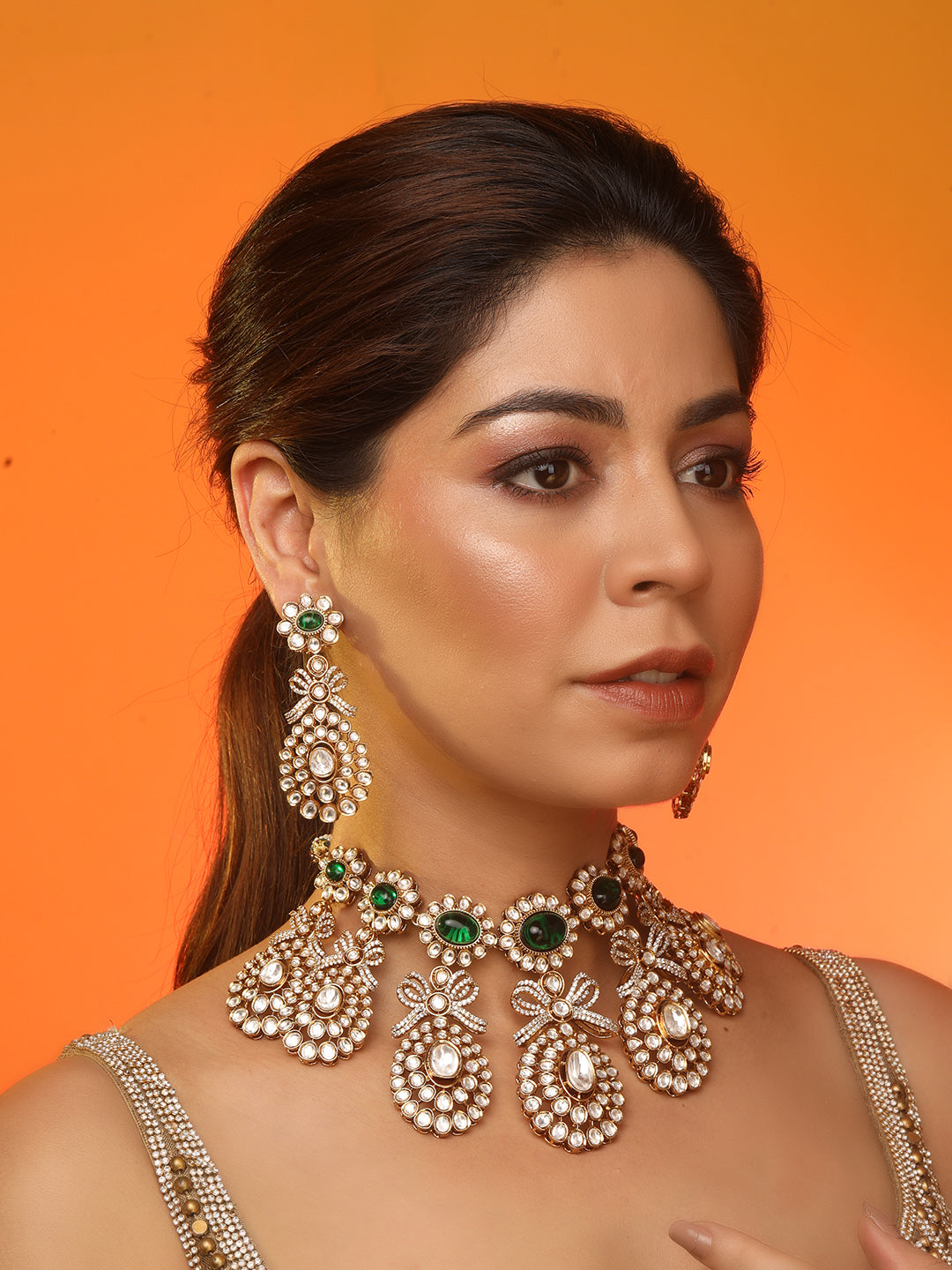 Gold-toned necklace with a braided cord and adjustable tassel tie, featuring intricate white stone detailing with oval green gemstones and multiple dangling teardrop motifs, paired with matching drop earrings, displayed on a plain light background.