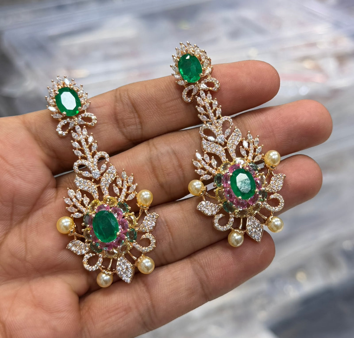 Rose gold plated emerald green American diamond long chandelier earrings with pink stones and pearl accents held in hand