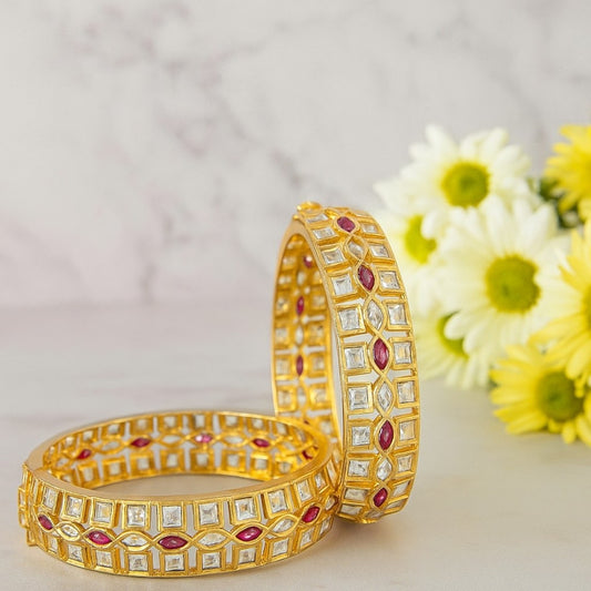 Elegant gold-plated bangles with square white stones and marquise ruby stones, placed on a marble background with yellow daisies – perfect for bridal and festive wear.