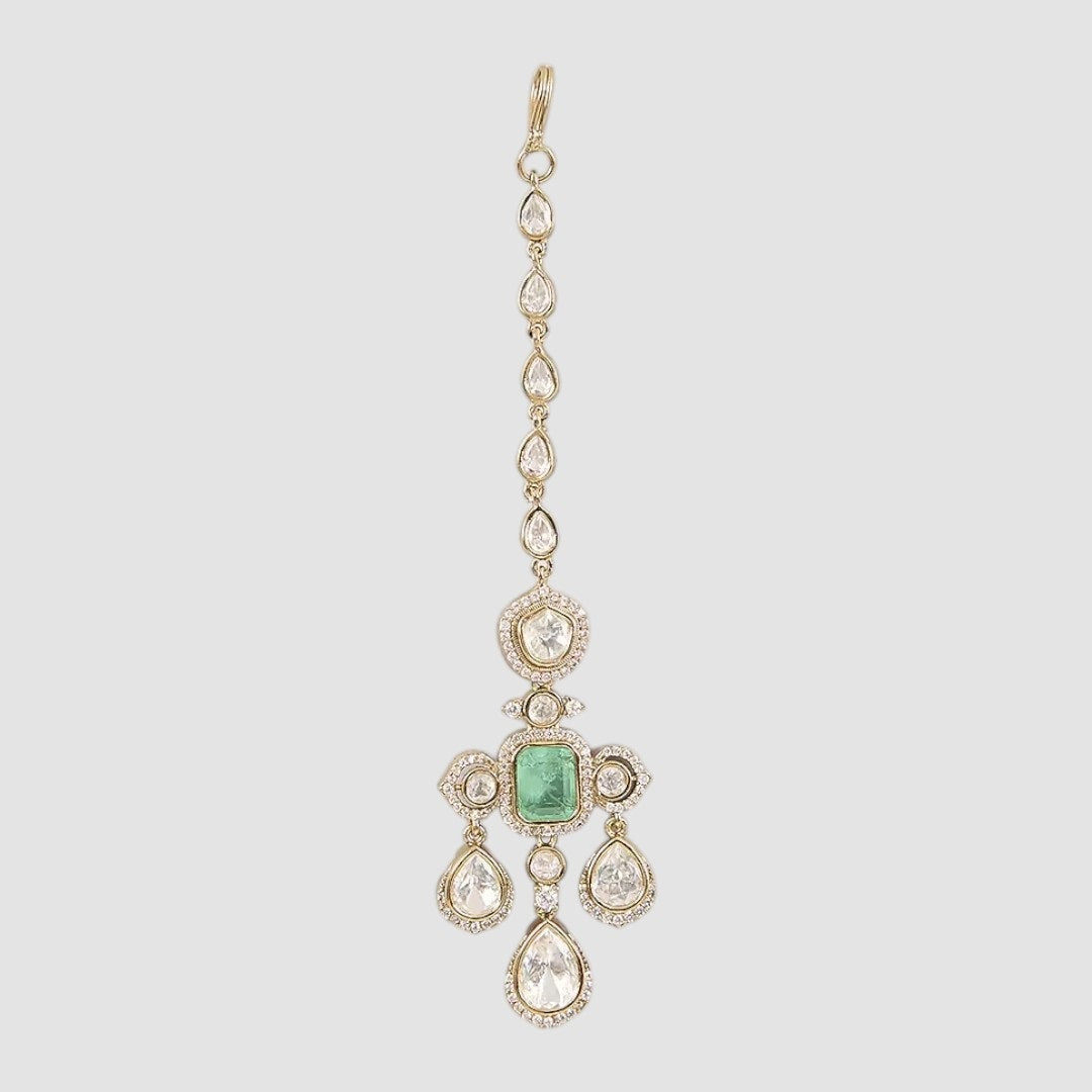 Gold-plated kundan maang tikka with emerald center and pear-shaped stones on a neutral background