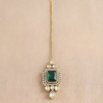 Gold maang tikka with emerald cut green stone in kundan halo setting and pearl chain, Indian bridal hair jewelry on white background