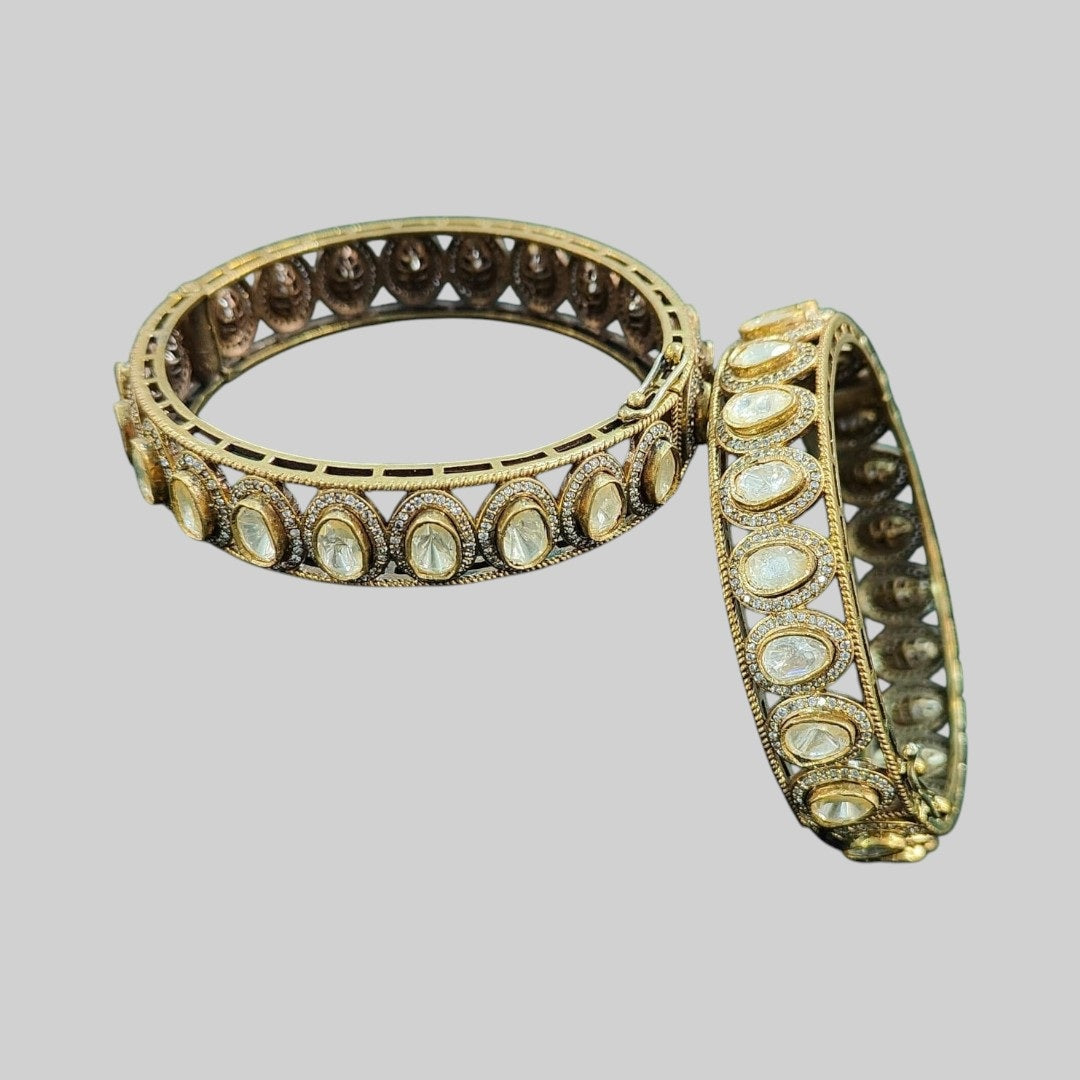 "Antique gold-plated bangles with oval polki stones in traditional Kundan style, placed on a green velvet cloth with floral background"