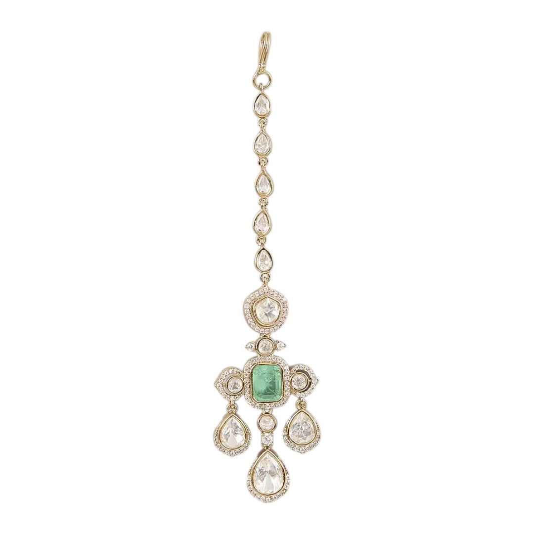 Gold-plated kundan maang tikka with emerald center and pear-shaped stones on a neutral background
