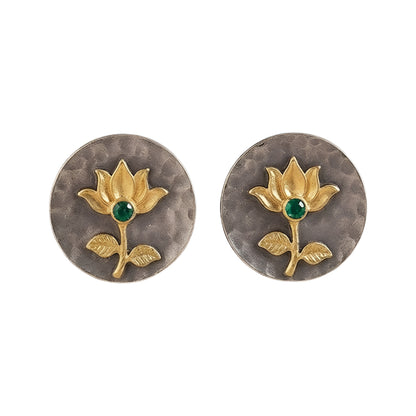 Gold lotus flower stud earrings with green emerald center on hammered oxidized silver disc, placed on a beige fabric background