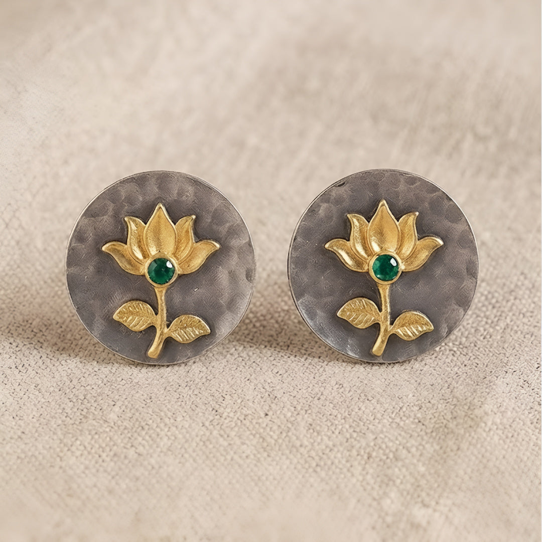 Gold lotus flower stud earrings with green emerald center on hammered oxidized silver disc, placed on a beige fabric background