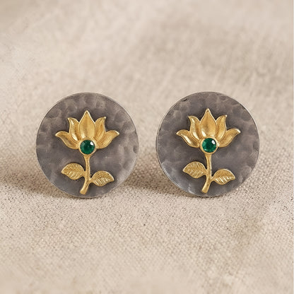 Gold lotus flower stud earrings with green emerald center on hammered oxidized silver disc, placed on a beige fabric background