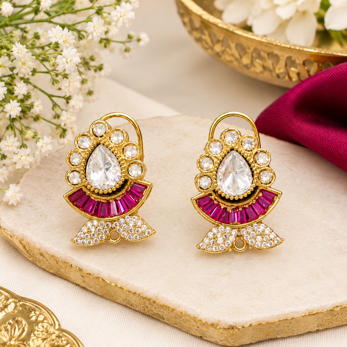 Pair of gold kundan-style stud earrings with a clear pear centre stone, ruby-pink baguette fan and zircon leaf base on a marble slab